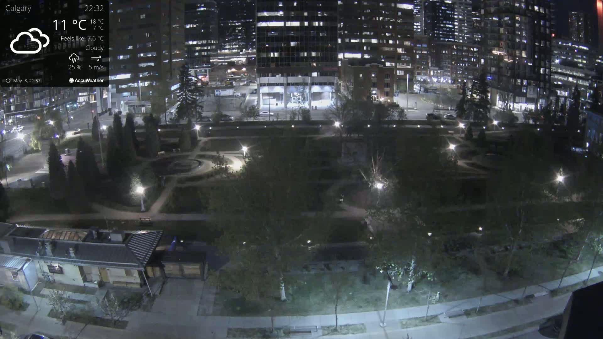A nighttime, cloudy view of a city park with illuminated pathways and trees, adjacent to brightly lit high-rise buildings.