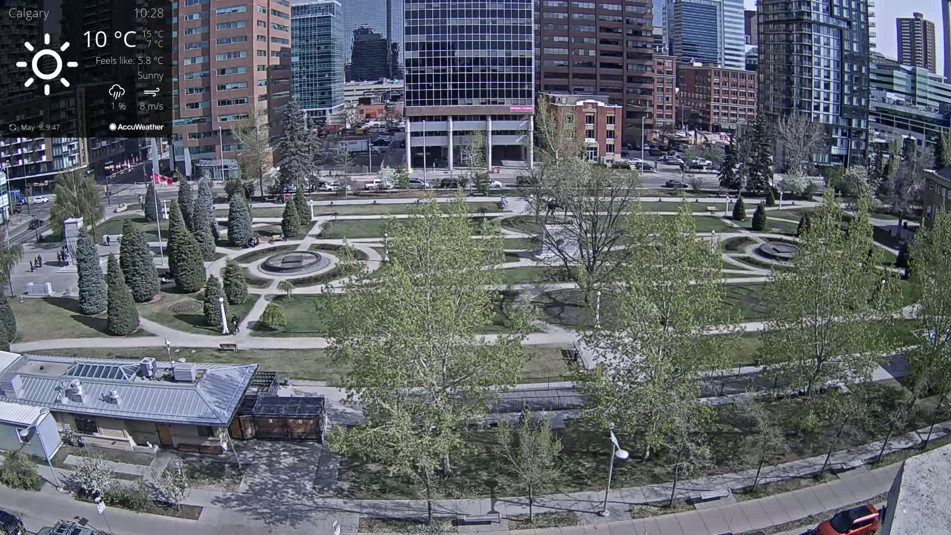 A sunny city park with green trees and paved pathways is viewed from above, surrounded by tall buildings.