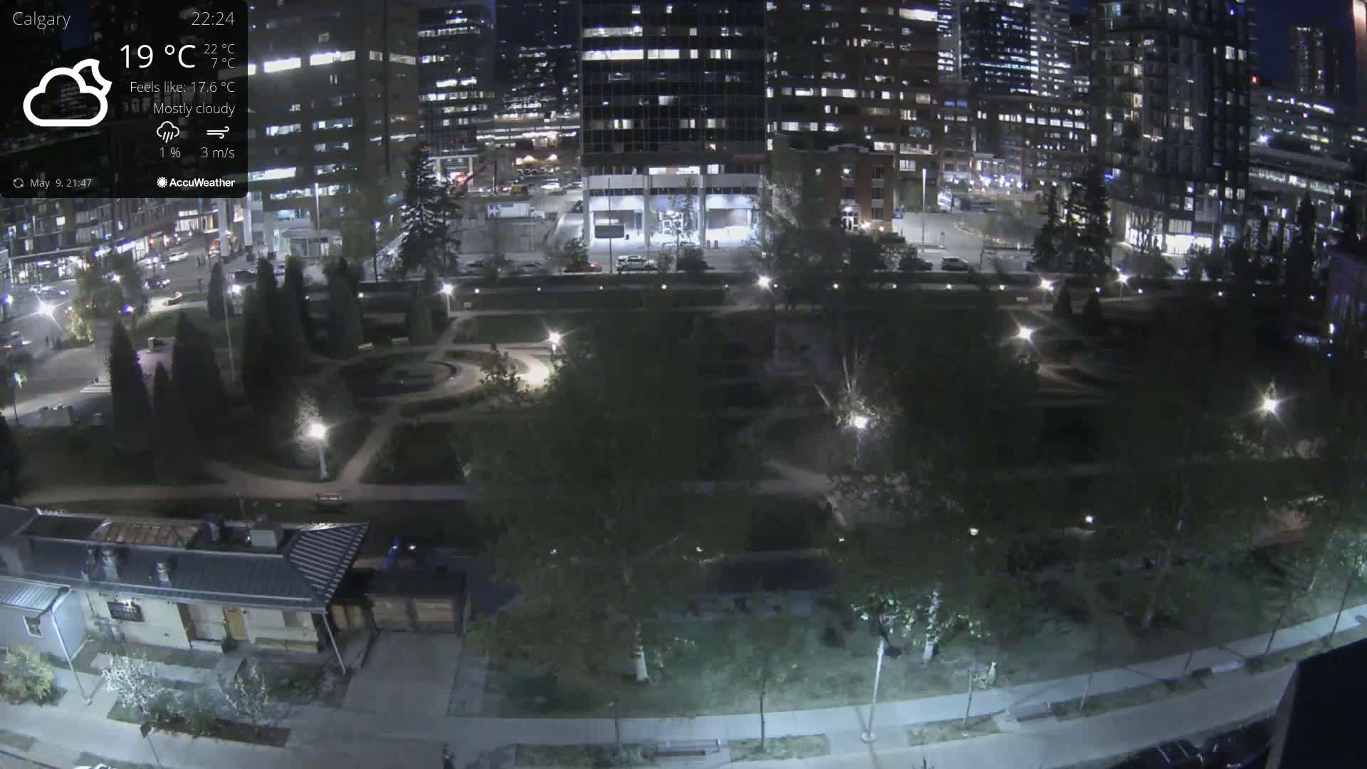 A mostly cloudy nighttime scene shows a park with pathways and trees, bordered by city buildings and streets with some vehicle lights.
