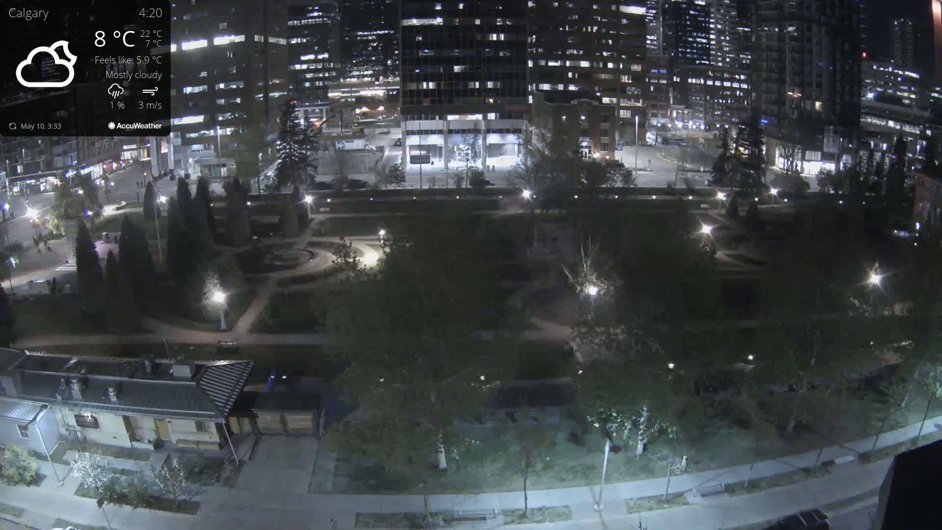 A mostly cloudy night scene shows a park with paved pathways, surrounded by buildings and illuminated by streetlights.