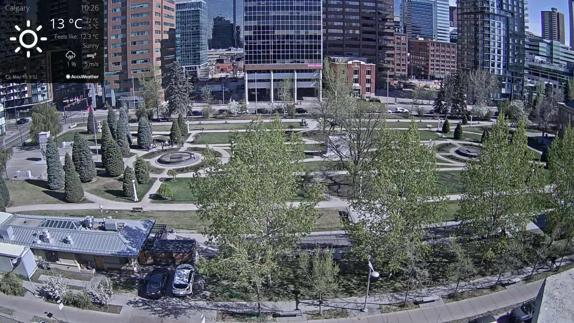 A sunny cityscape shows a park with trees and paved walkways, next to buildings under a clear sky.