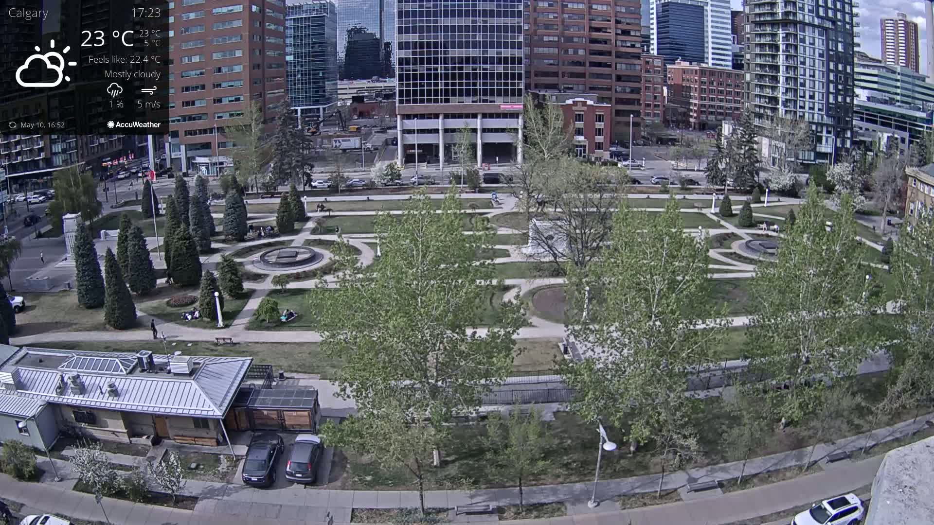 A mostly cloudy city park with a fountain and several people scattered throughout, surrounded by buildings and trees.