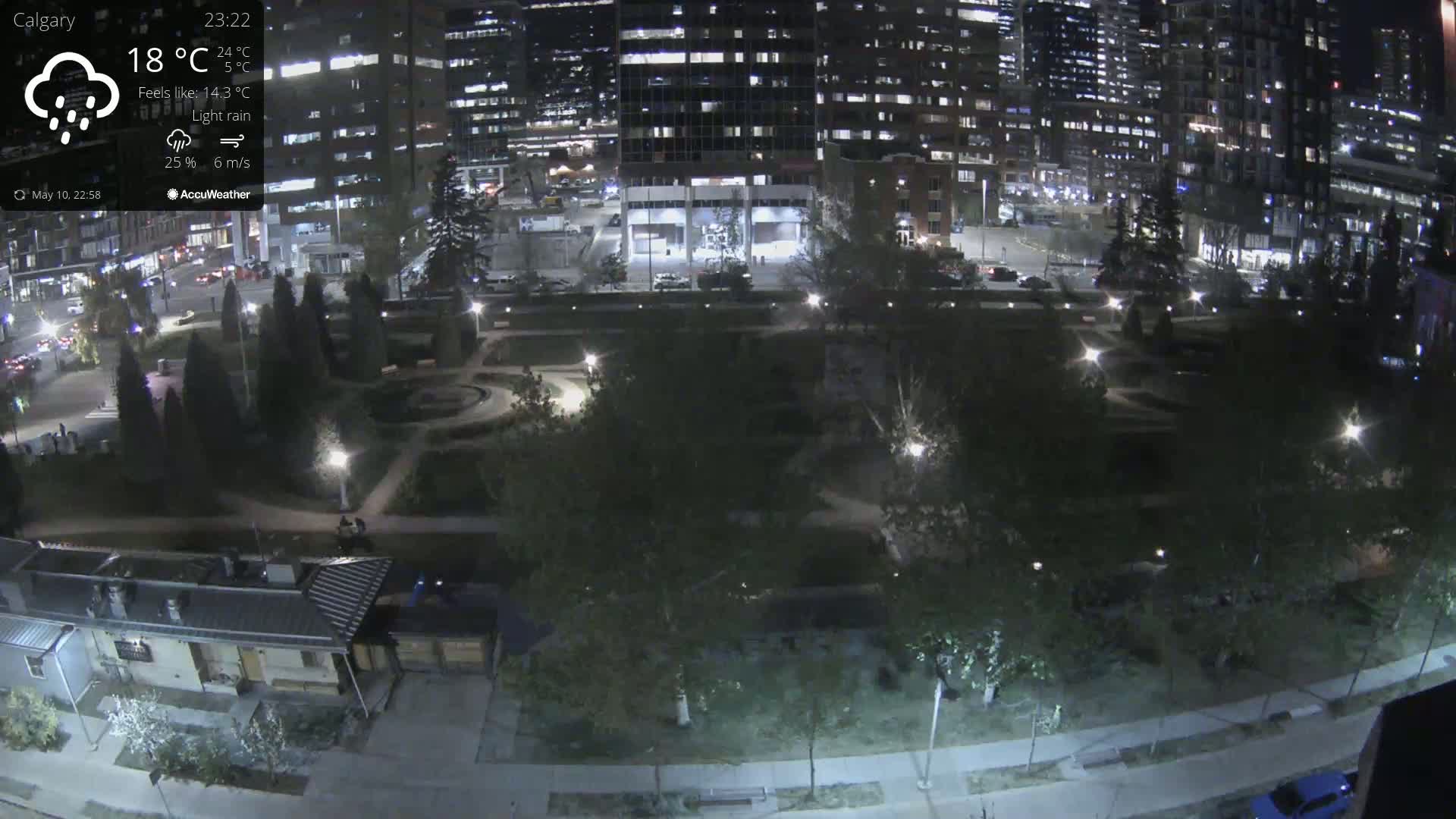 An aerial nighttime view of a city park, lightly rain-sprinkled, with city buildings illuminated in the background.