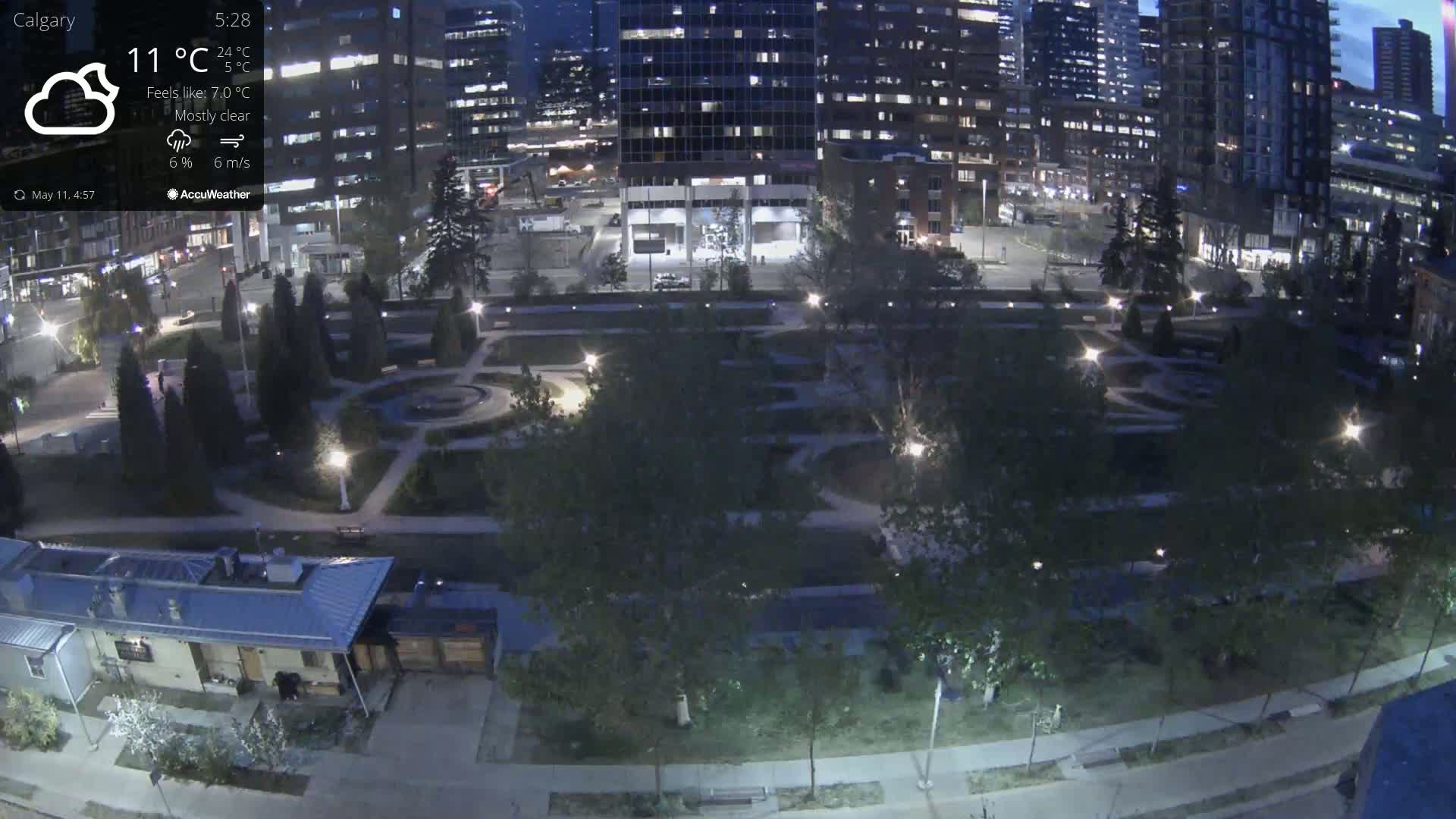 A mostly clear night scene shows a city park illuminated by lights, bordered by tall buildings.