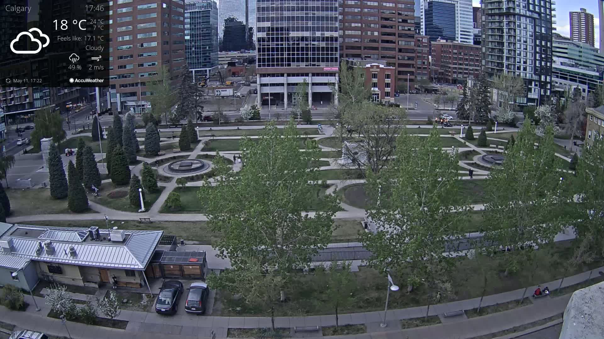 A cloudy urban park with a fountain, surrounded by trees and buildings, is viewed from above on a cool day.