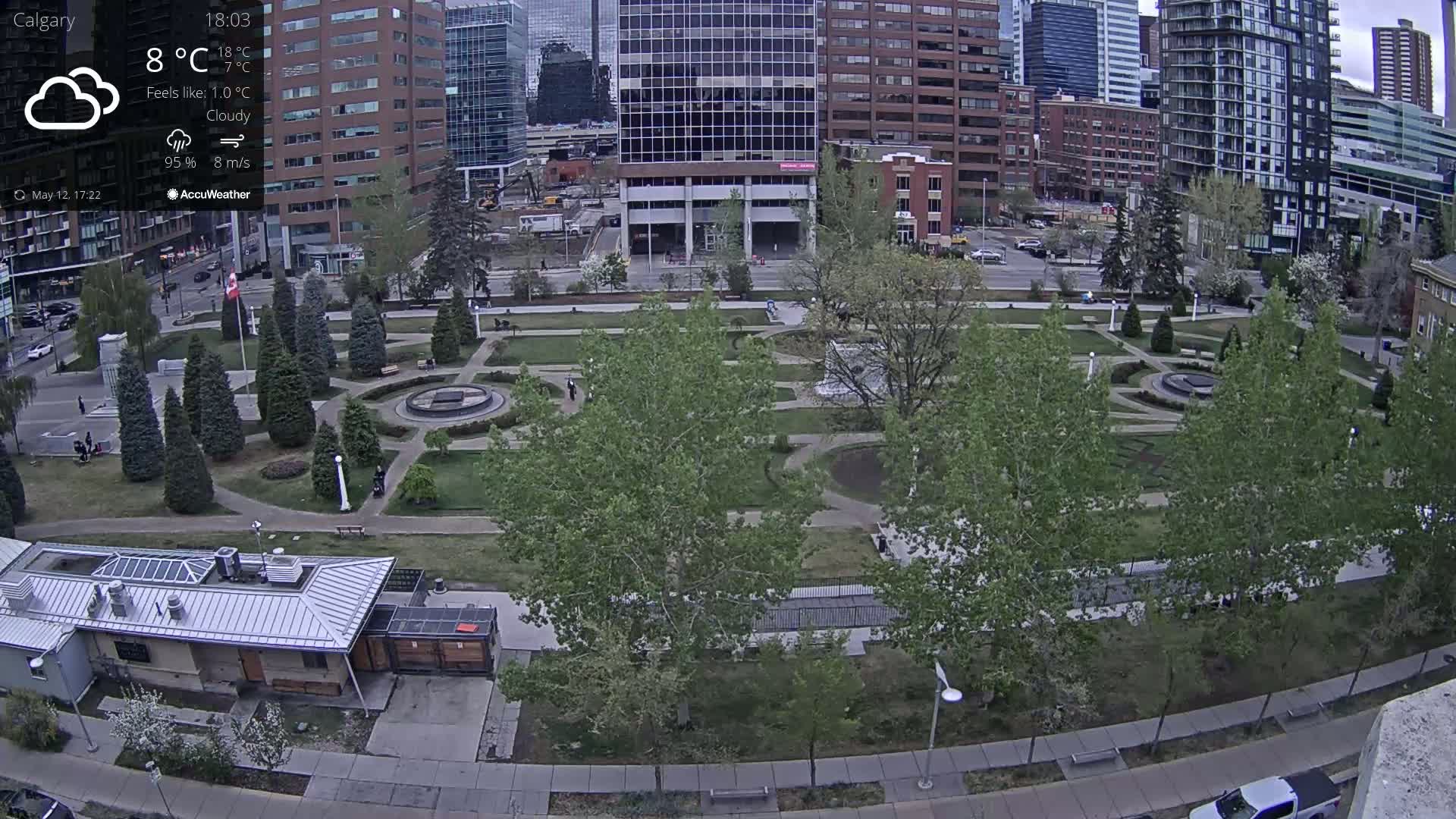 A city park with lush green landscaping and numerous trees is situated between tall buildings under cloudy skies.