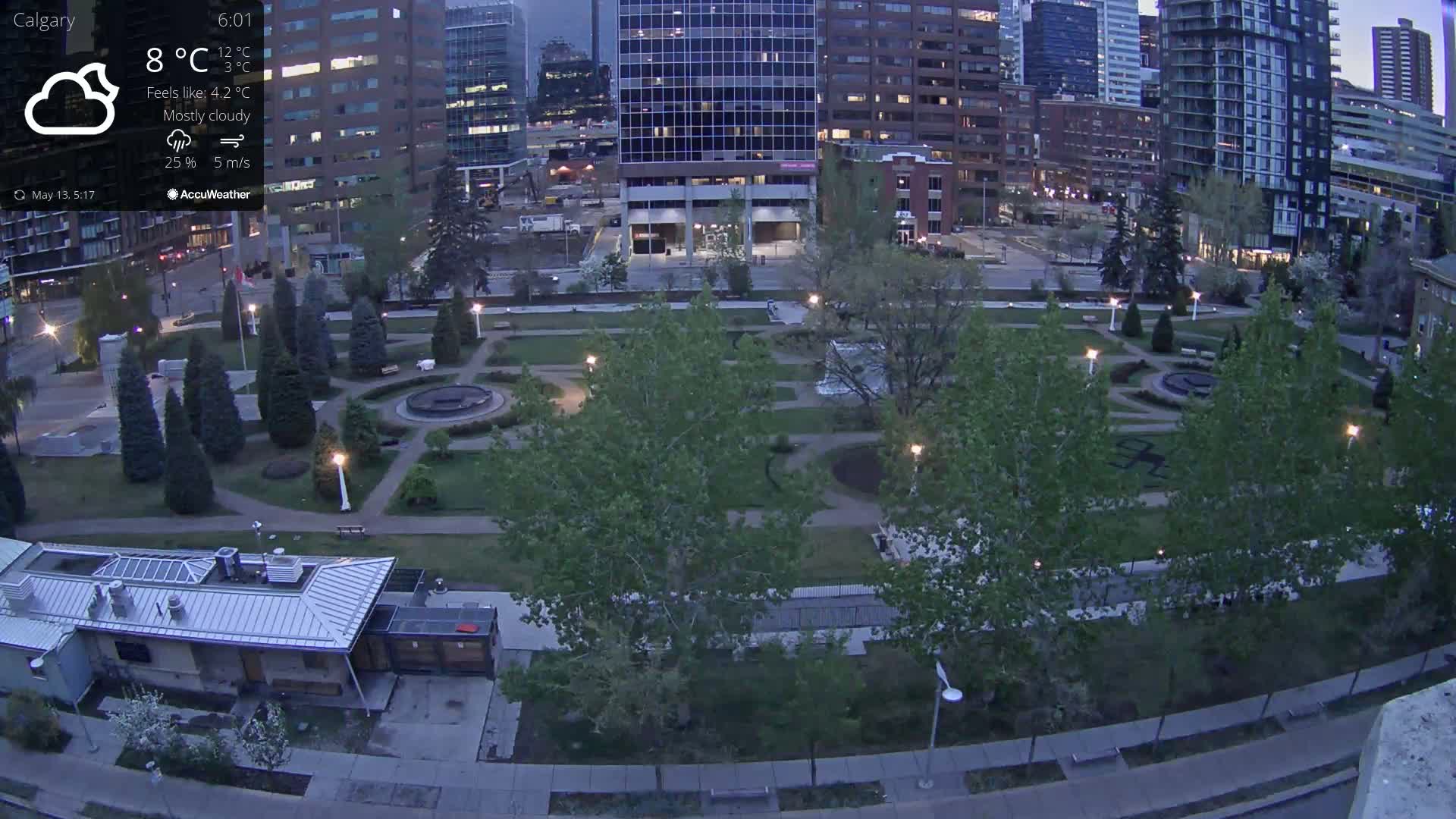 A mostly cloudy evening scene shows a city park with green lawns, trees, and pathways, surrounded by tall buildings.