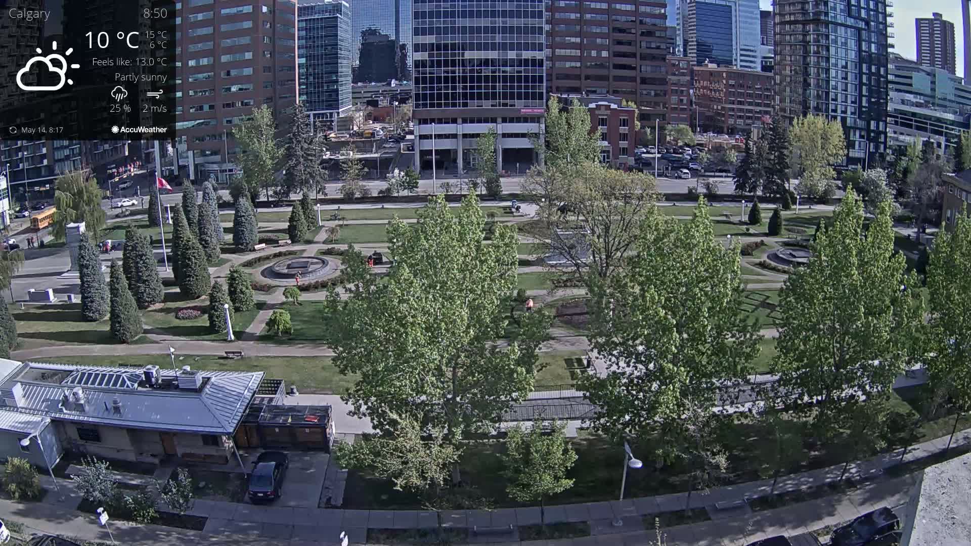 A partly sunny city park with green trees and a central fountain is surrounded by modern buildings, under a partly sunny sky.