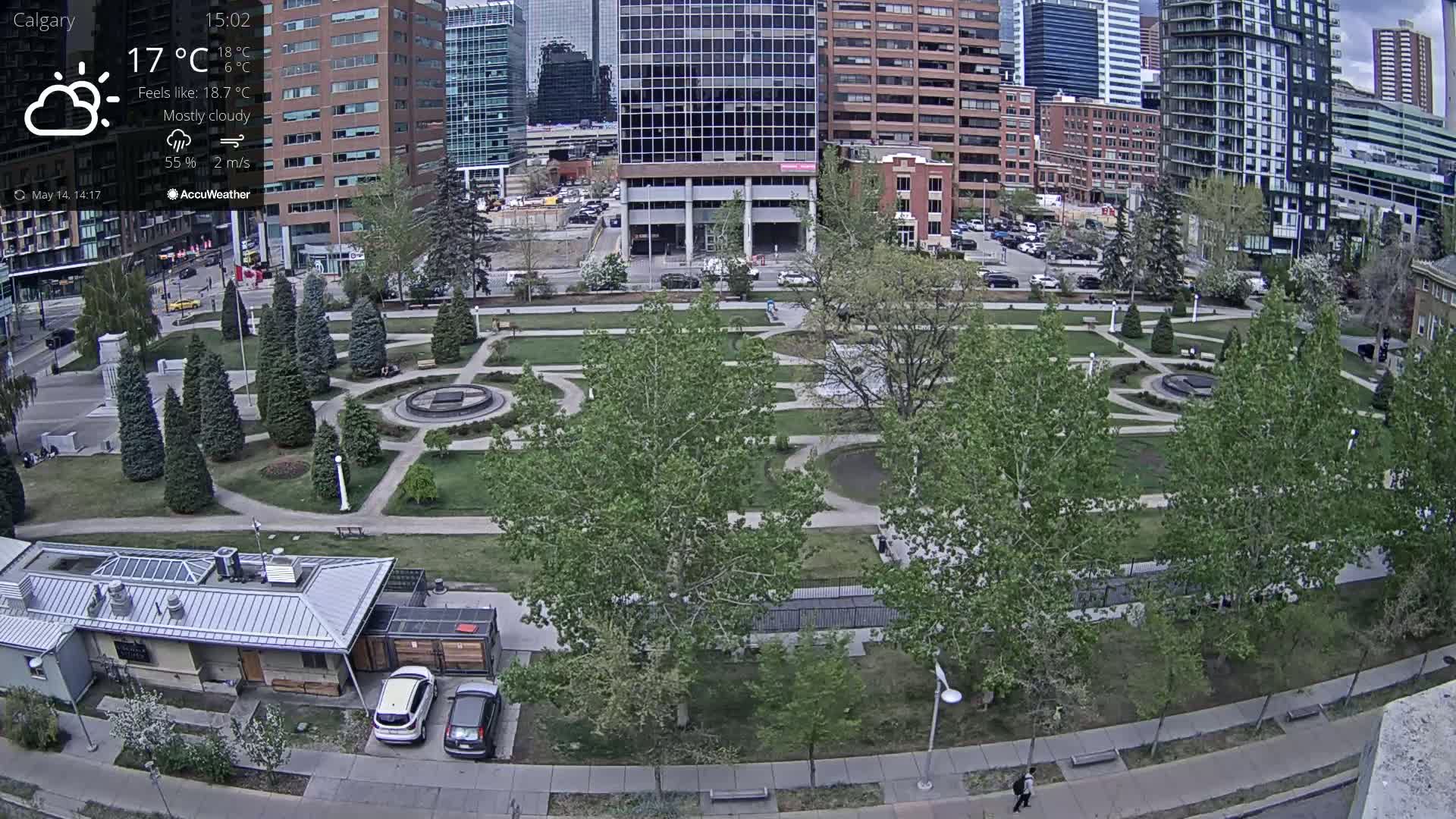 A mostly cloudy city park with green trees and shrubs, a fountain, and walkways, situated between modern and older buildings.