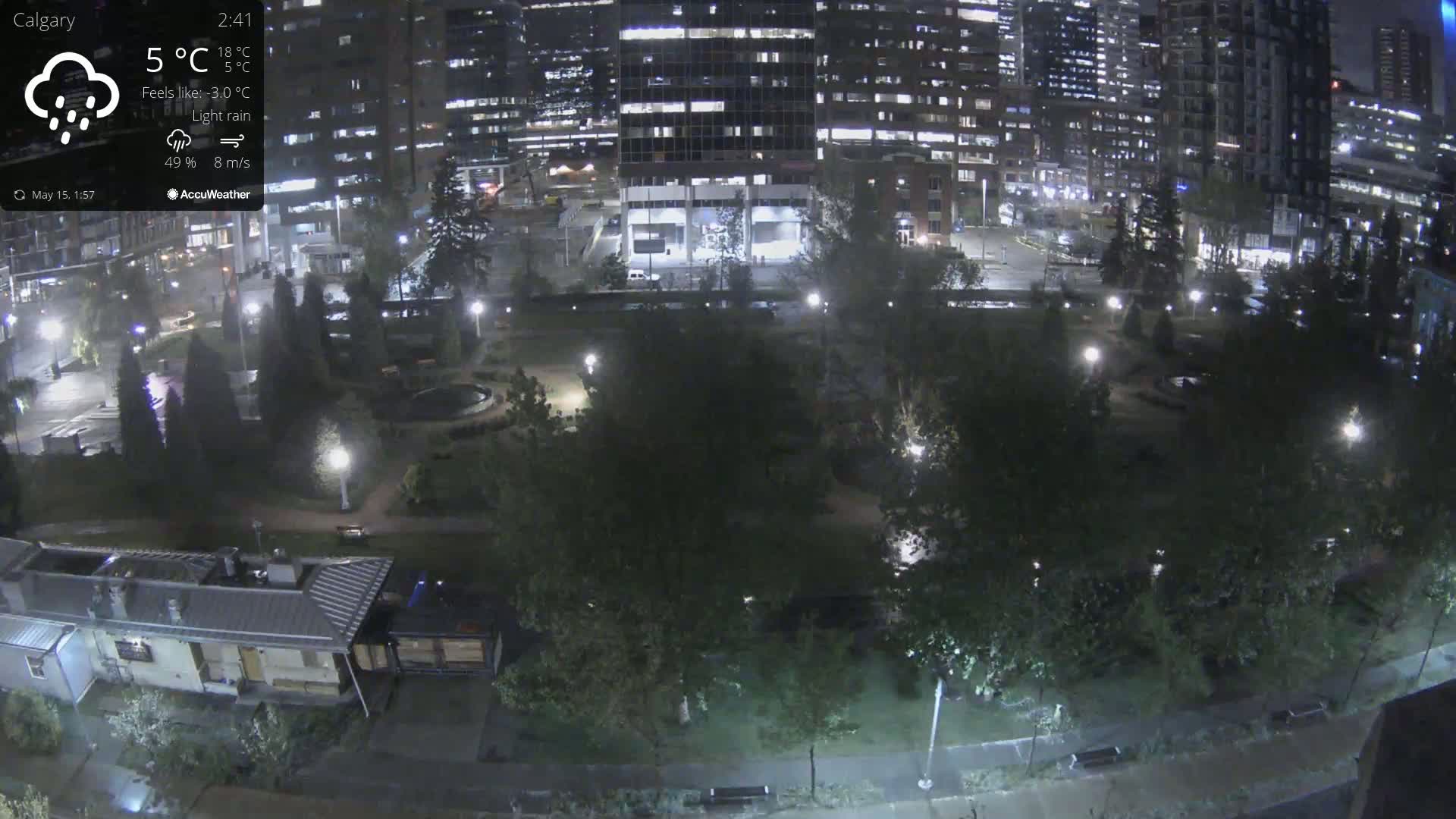 A nighttime view of a park with light rain, illuminated by streetlights, adjacent to brightly lit city buildings.