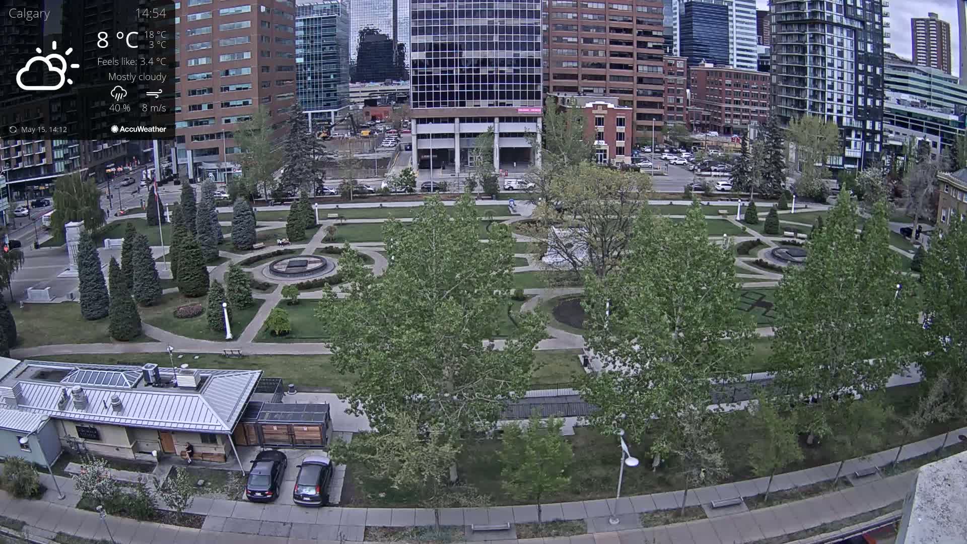 An aerial view of a city park on a mostly cloudy day, with eight-meter-per-second winds, showing green lawns, paved walkways, trees, and a few cars parked near a small building.