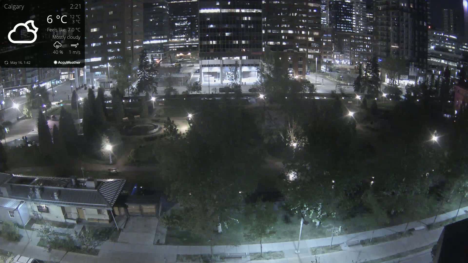 A nighttime, mostly cloudy view of a city park bordered by illuminated buildings and trees.