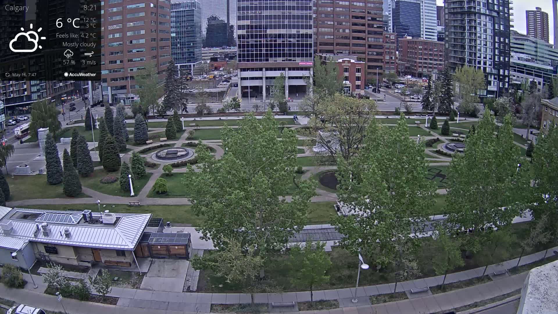 A mostly cloudy, cool day overlooks a city park with a fountain and several mature trees, adjacent to modern and older buildings.