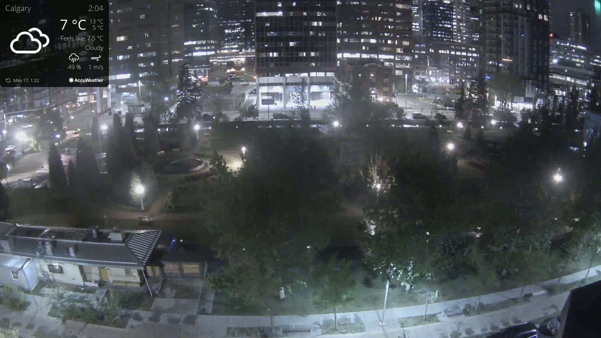 A nighttime, cloudy view of a city park surrounded by illuminated buildings, under a light mist or fog.