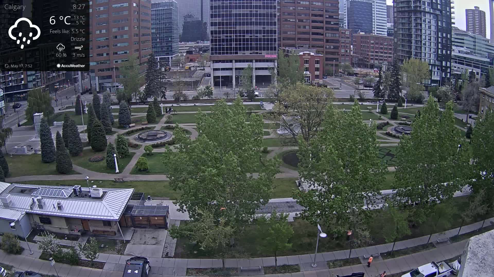 A city park with manicured lawns, trees, and fountains is seen under a drizzly sky, with city buildings in the background.