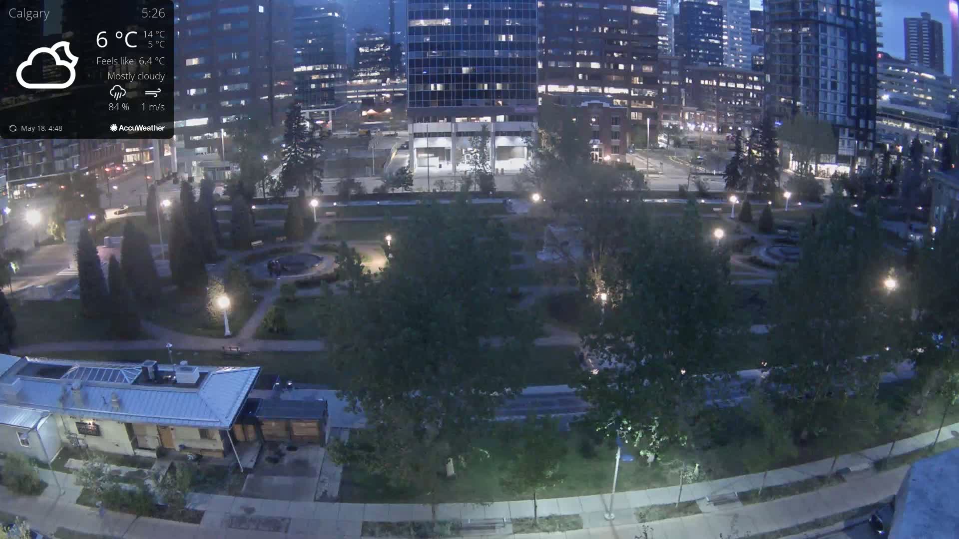 A mostly cloudy night scene shows a city park with trees and walkways, surrounded by tall buildings with lights on, under dim nighttime lighting.