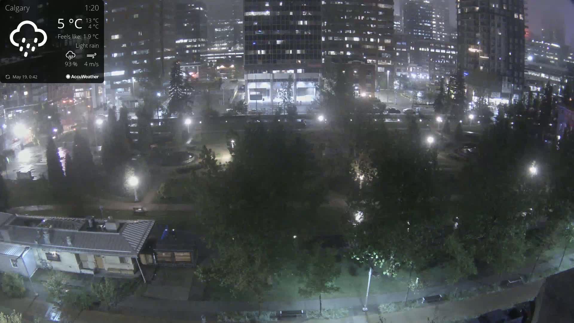 A nighttime view of a park surrounded by illuminated buildings under light rain.