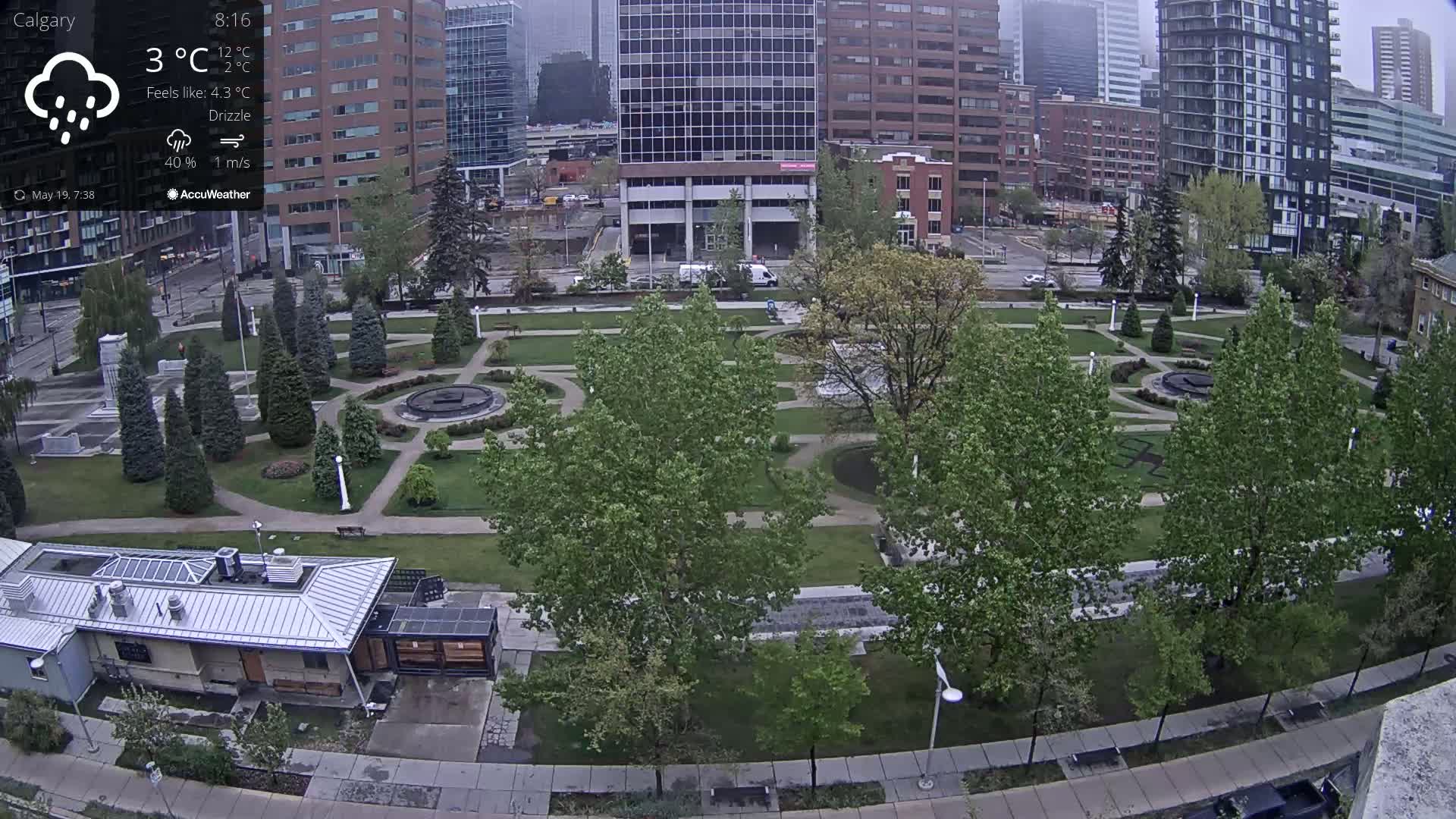 A city park with a fountain and several trees is shown under a light drizzle.