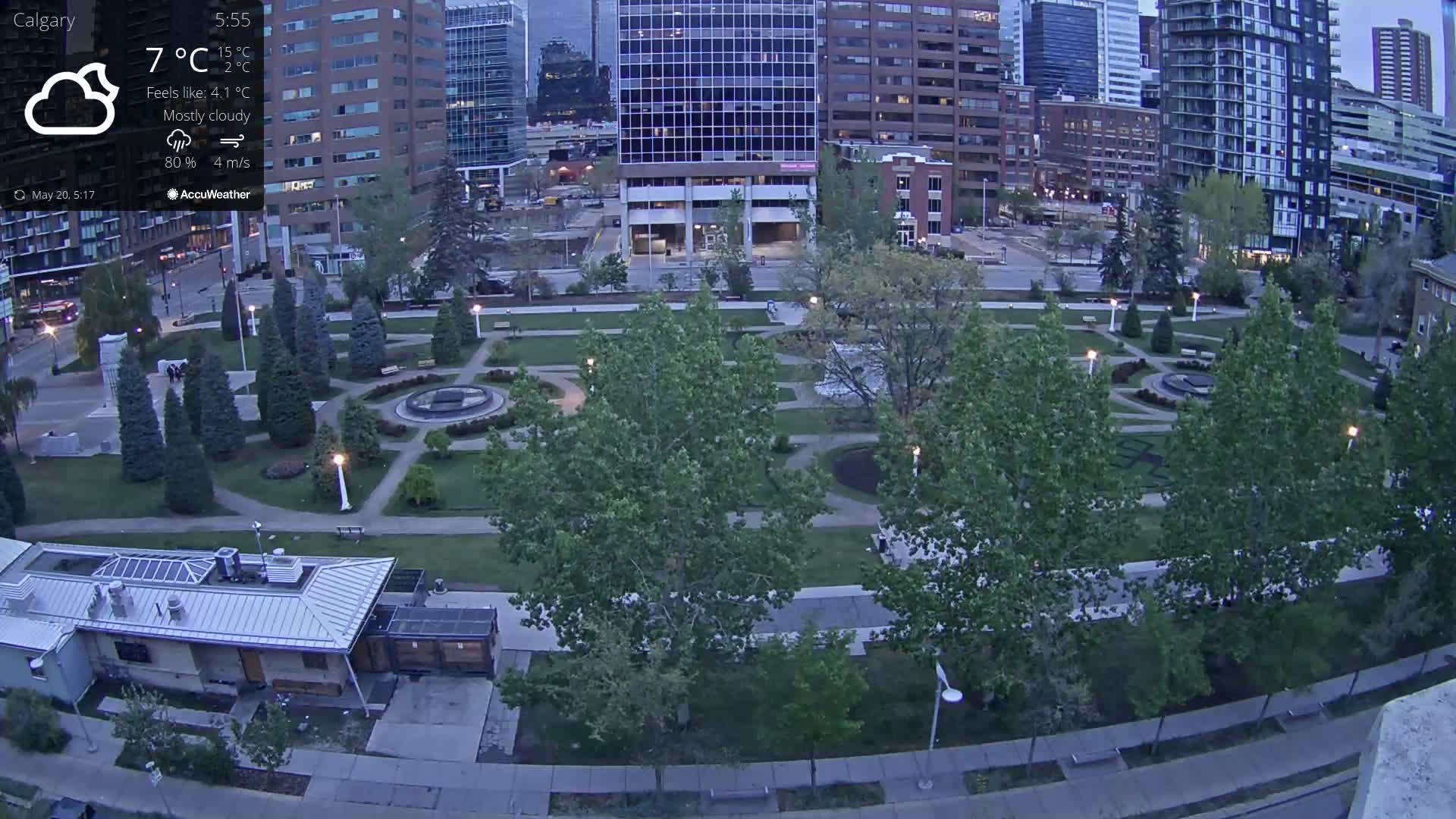 A mostly cloudy evening scene shows a city park with green trees and paved walkways, adjacent to tall city buildings.