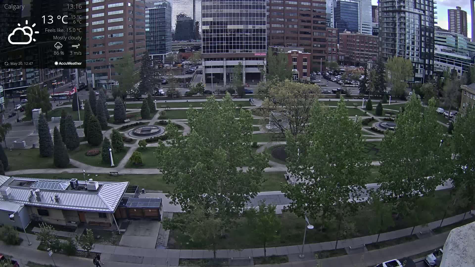A mostly cloudy city park with a fountain, surrounded by buildings and trees, on a 15°C day.