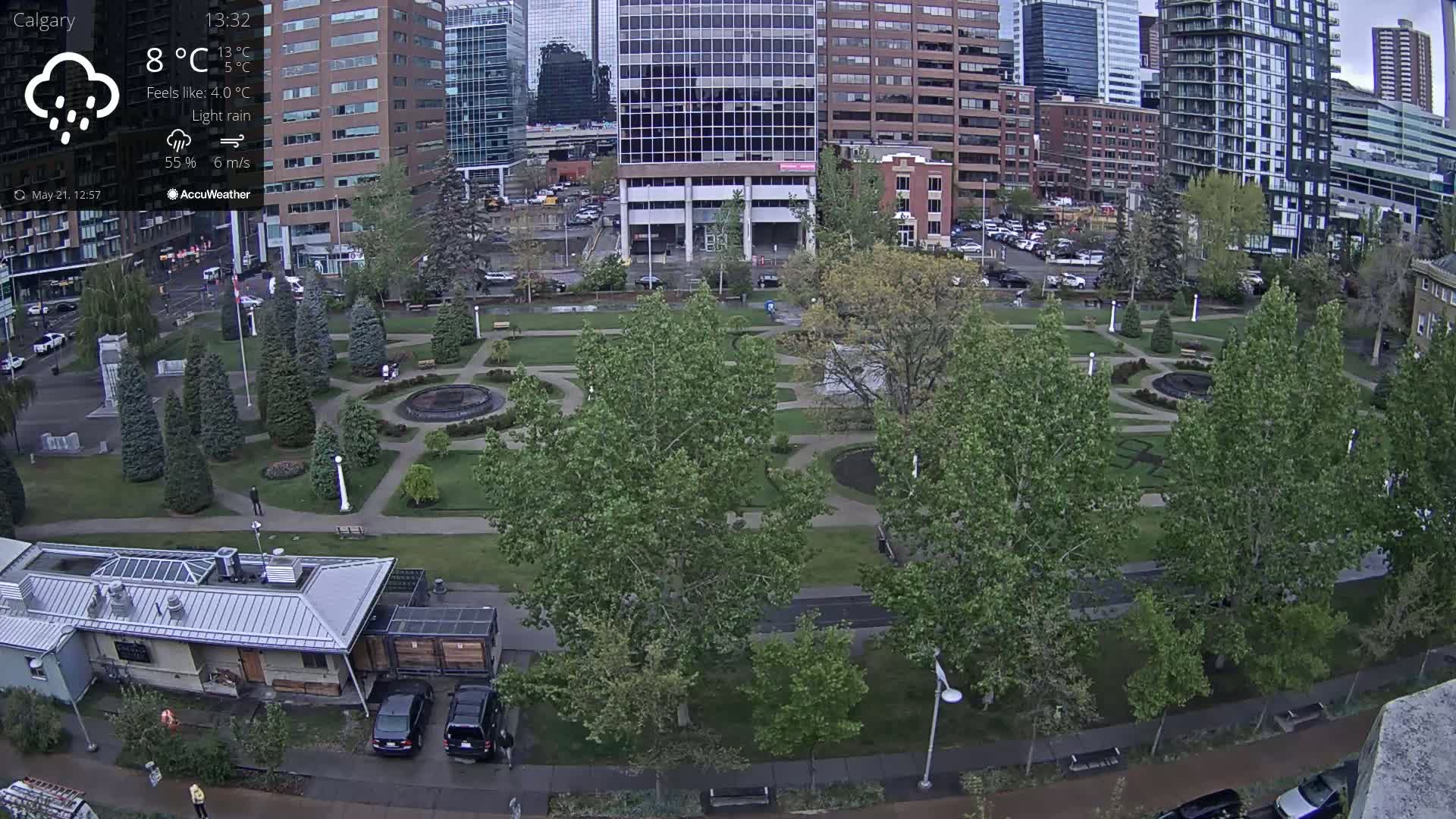 A city park with a fountain and landscaping is seen under light rain, surrounded by tall buildings.