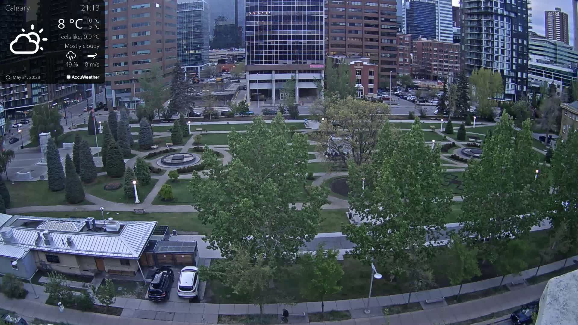 An aerial view of a city park with mostly cloudy skies, under which are green trees and paved walkways, two cars parked near a building, and city buildings in the background.