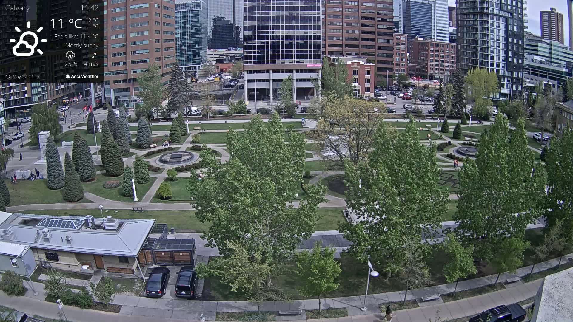 A mostly sunny urban park with a central fountain, surrounded by lush green trees and paved walkways, sits between modern high-rise buildings.