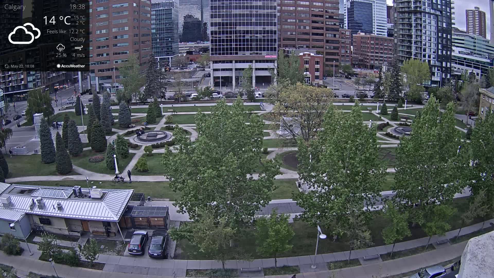 An aerial view of a city park on a cloudy day, showing manicured lawns, walkways, trees, and buildings in the background.