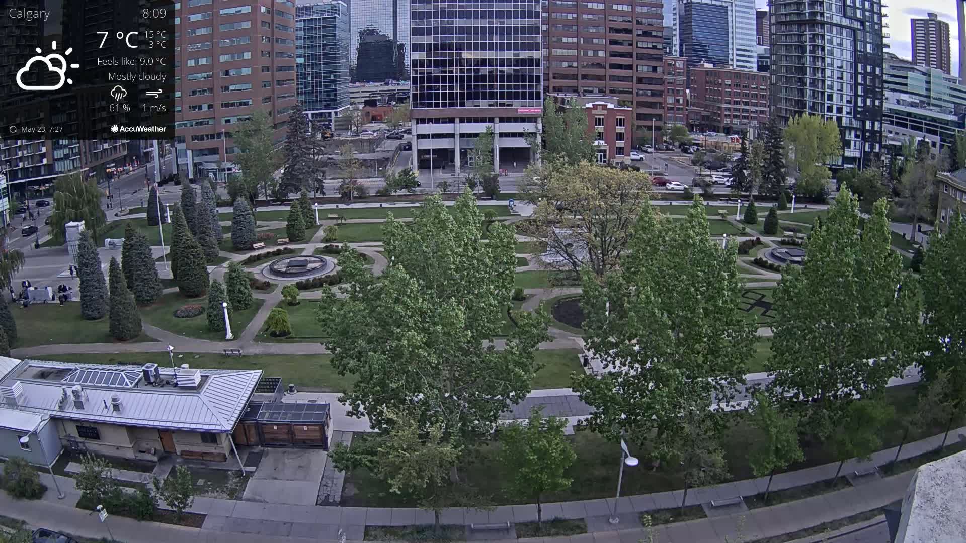 A mostly cloudy, 7°C day overlooks a city park with green trees and paved pathways, adjacent to tall buildings.