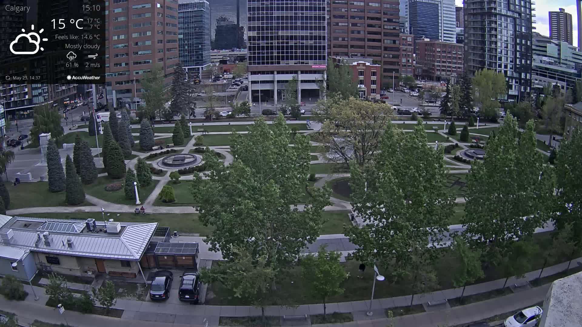 A mostly cloudy day reveals a city park with manicured lawns, paved pathways, and several mature trees, surrounded by tall buildings.