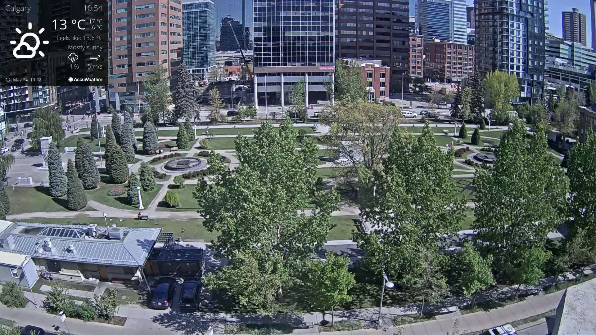 A mostly sunny city park with lush green trees and a central fountain is surrounded by modern high-rise buildings.