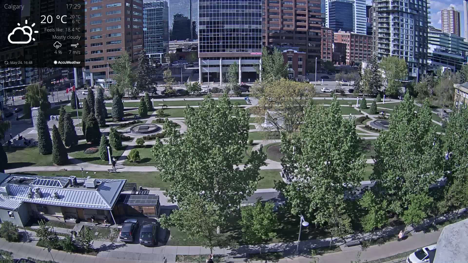 A mostly cloudy, sunny day reveals a park with many trees and paved walkways, situated between several city buildings.
