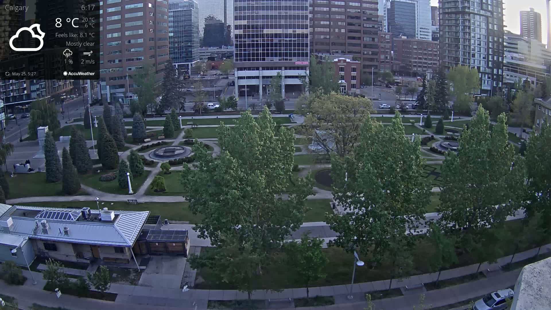 An aerial view of a city park with a fountain and several trees, under mostly clear skies.