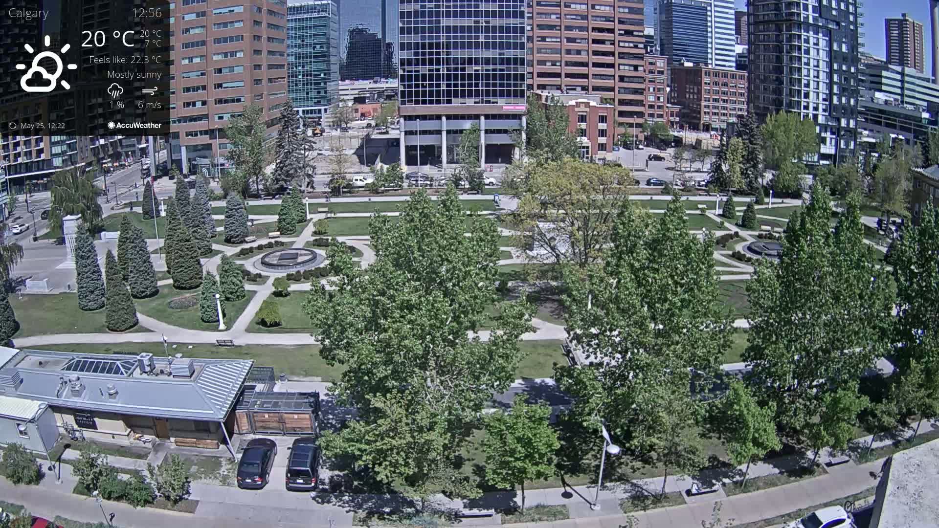 A mostly sunny city park with a fountain, surrounded by buildings and numerous green trees, under a mostly sunny sky.