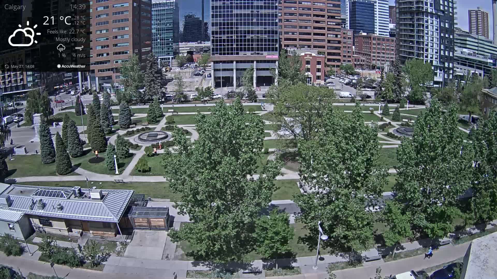 A mostly cloudy day with a temperature of 21°C shows a city park with lush green landscaping, trees, and walkways surrounded by modern buildings.