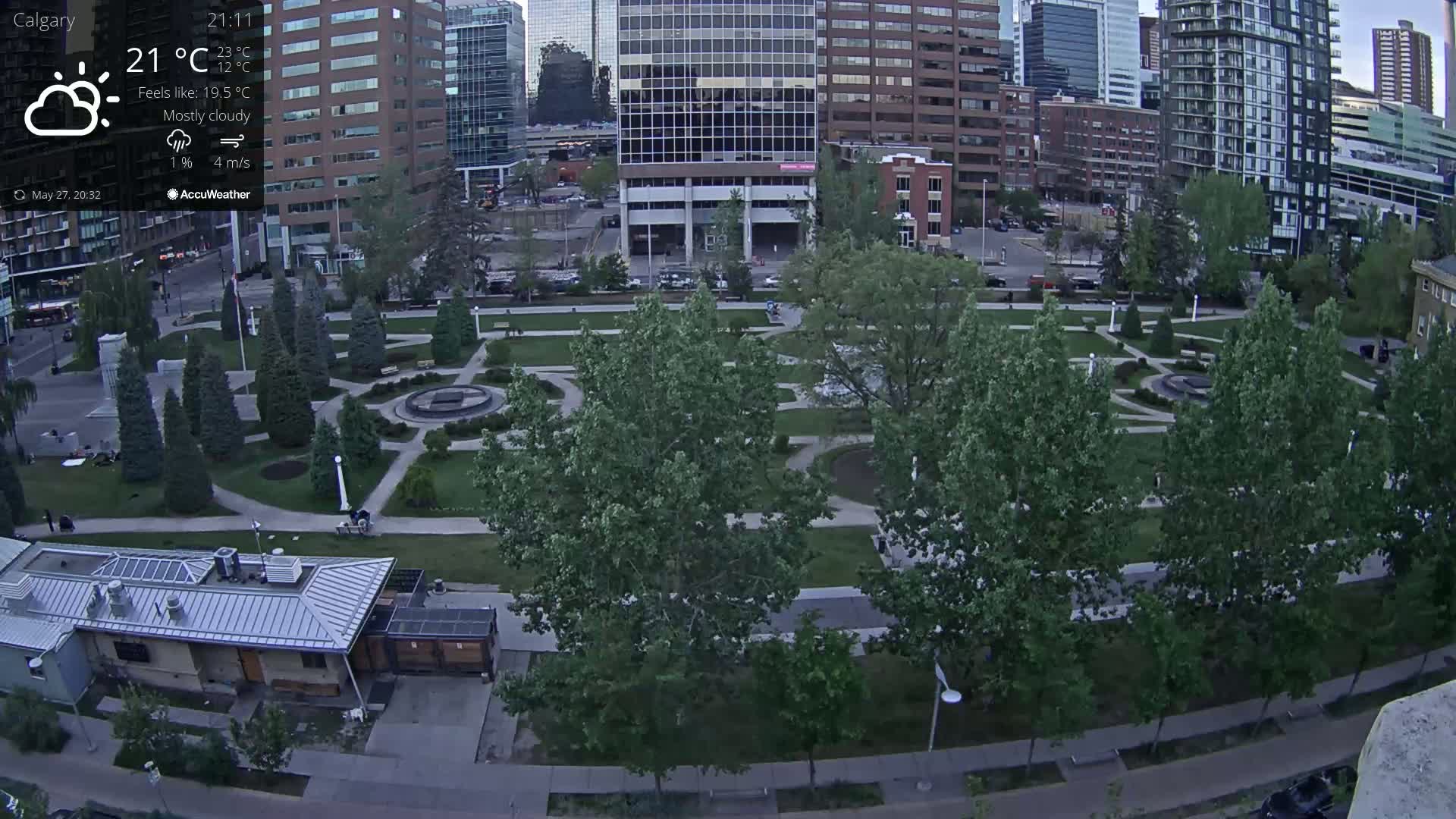 A mostly cloudy evening scene shows a city park with green trees and paved pathways, bordered by tall buildings.