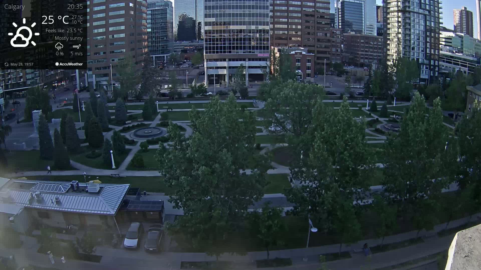 A mostly sunny, evening view of a city park with a fountain, surrounded by green trees and buildings.