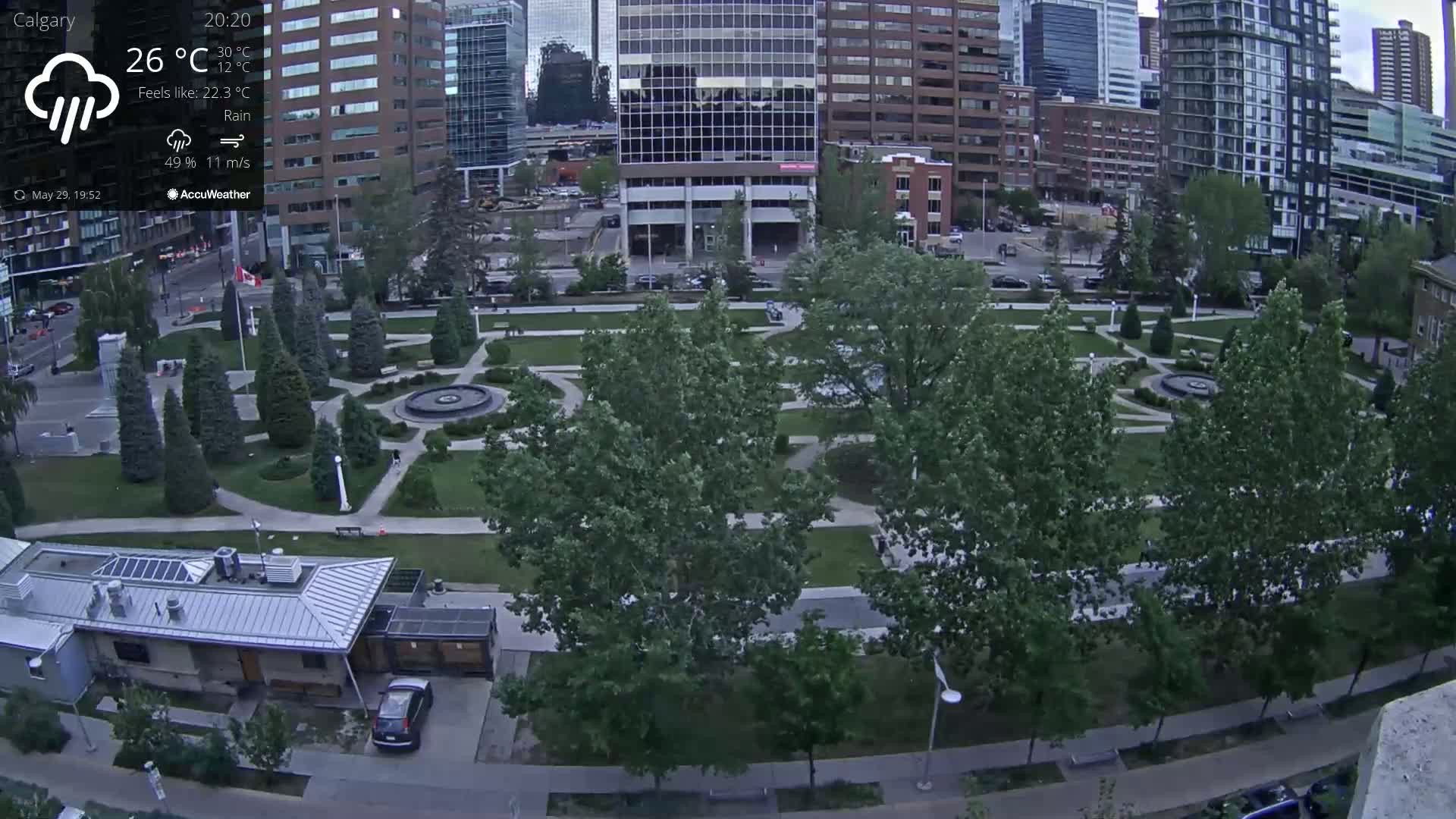 A city park with a fountain, surrounded by lush green trees and paved walkways, is seen under rainy conditions.