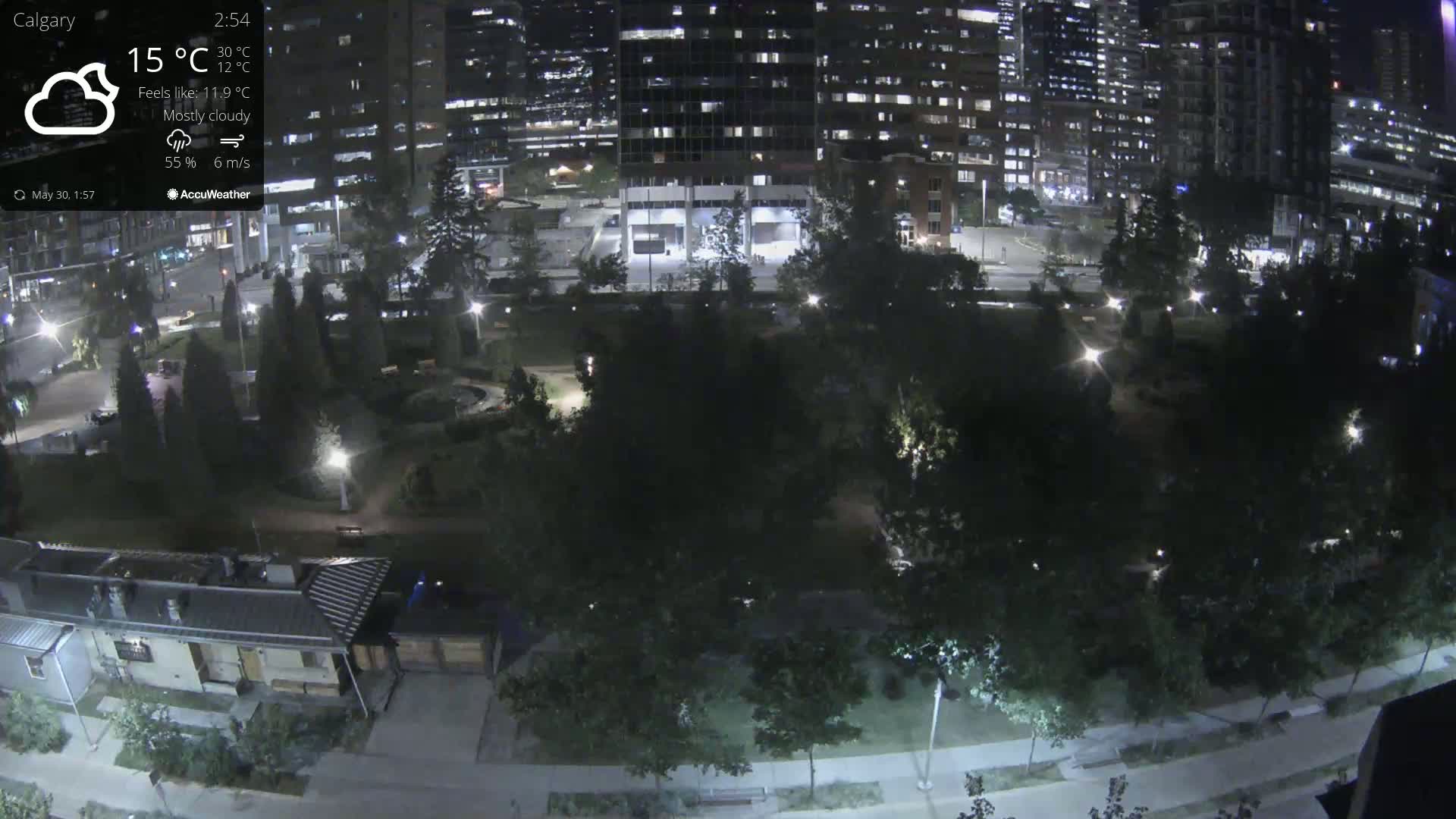 A mostly cloudy night scene shows a park with trees and pathways, surrounded by illuminated city buildings.