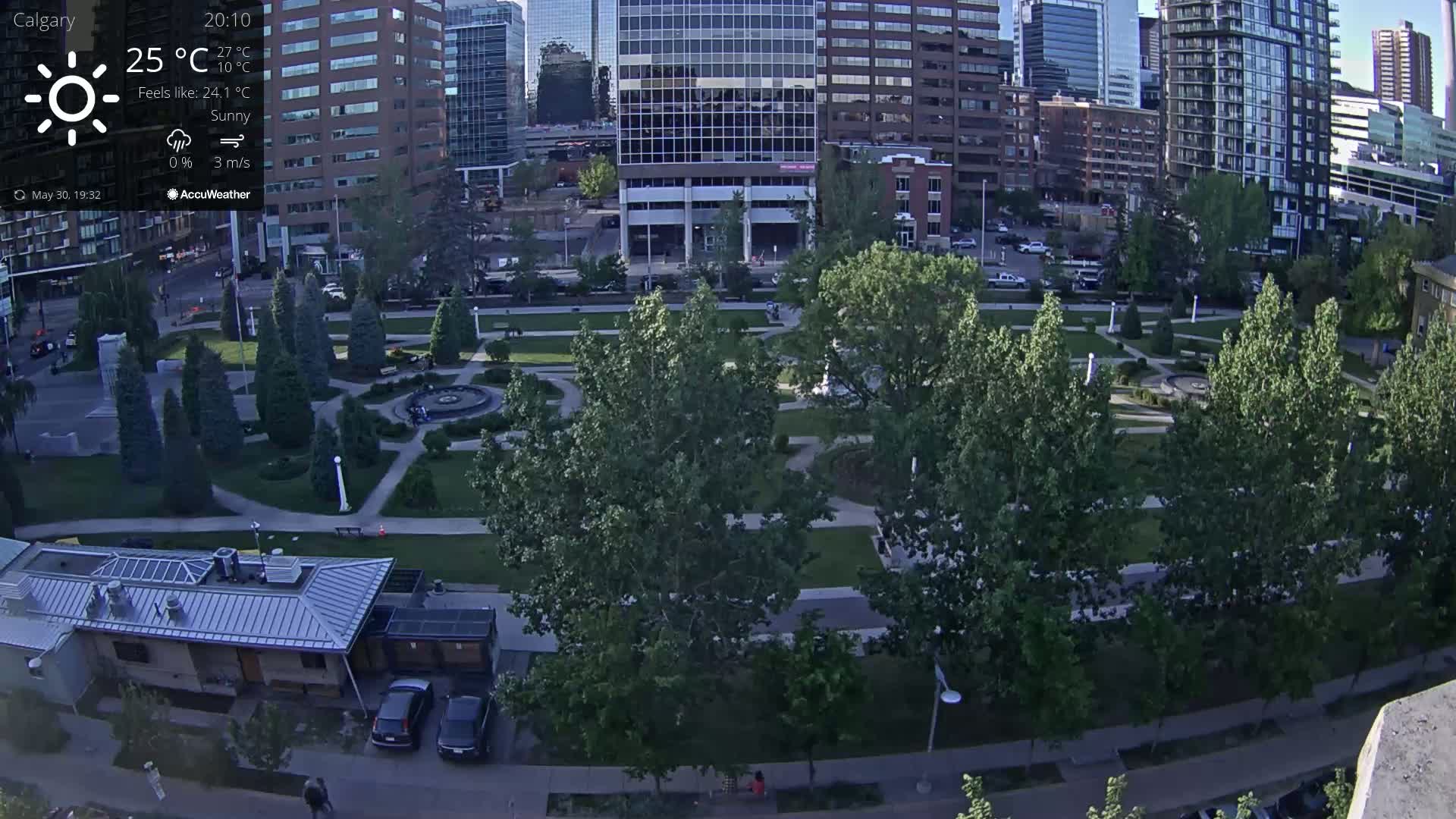 A sunny cityscape with a park containing several trees and a fountain is viewed from above.