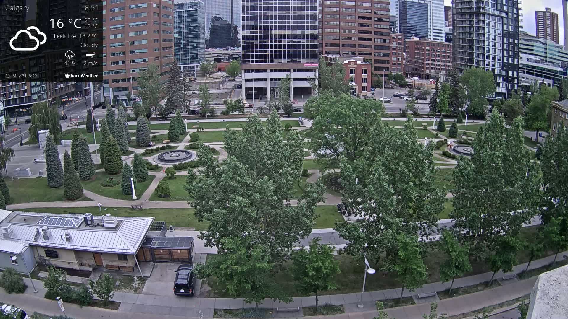 A cloudy city park with a fountain, surrounded by tall buildings, under a partly cloudy sky.