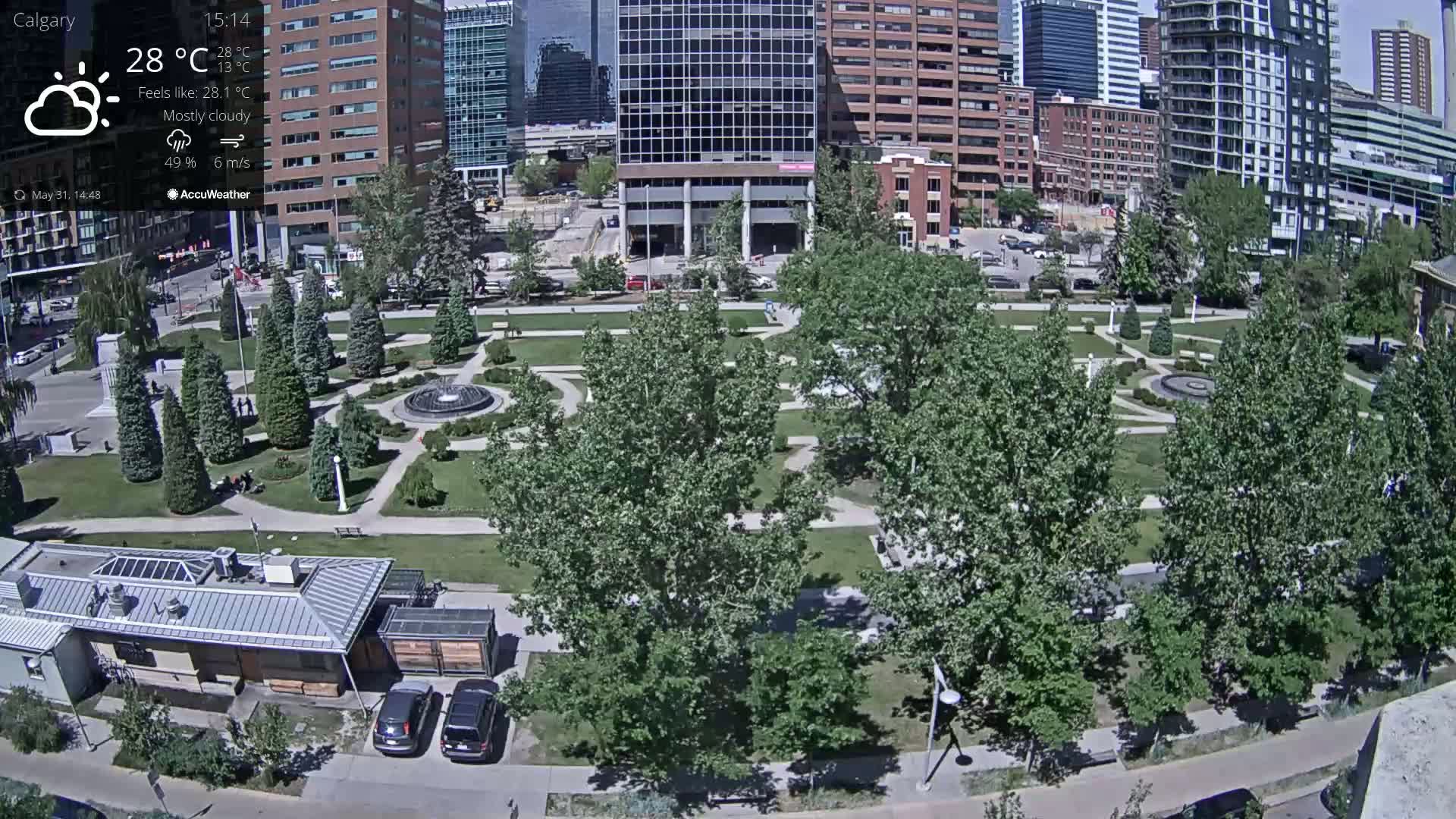 A mostly cloudy, 28°C day overlooks a city park with a fountain, several trees, and buildings in the background.