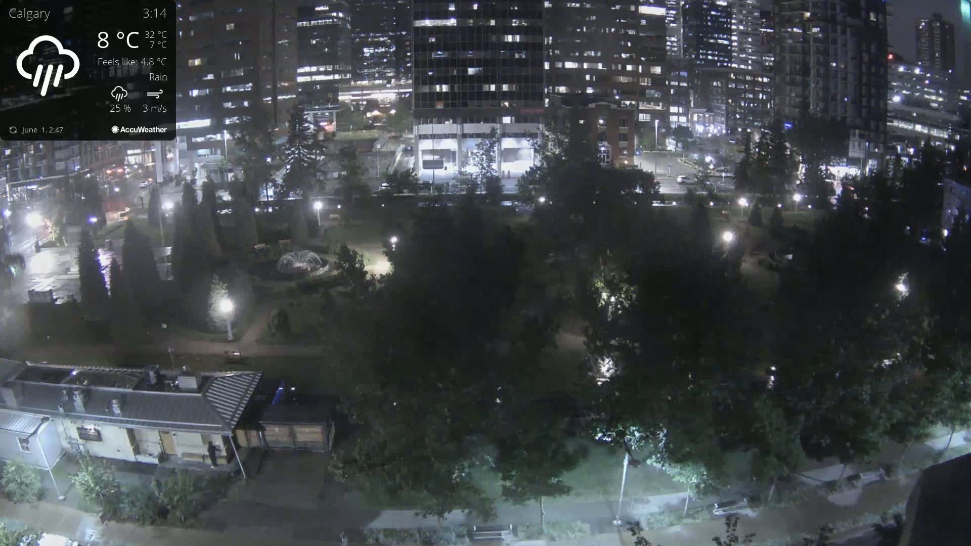 A nighttime, rainy view of a city park surrounded by illuminated buildings.