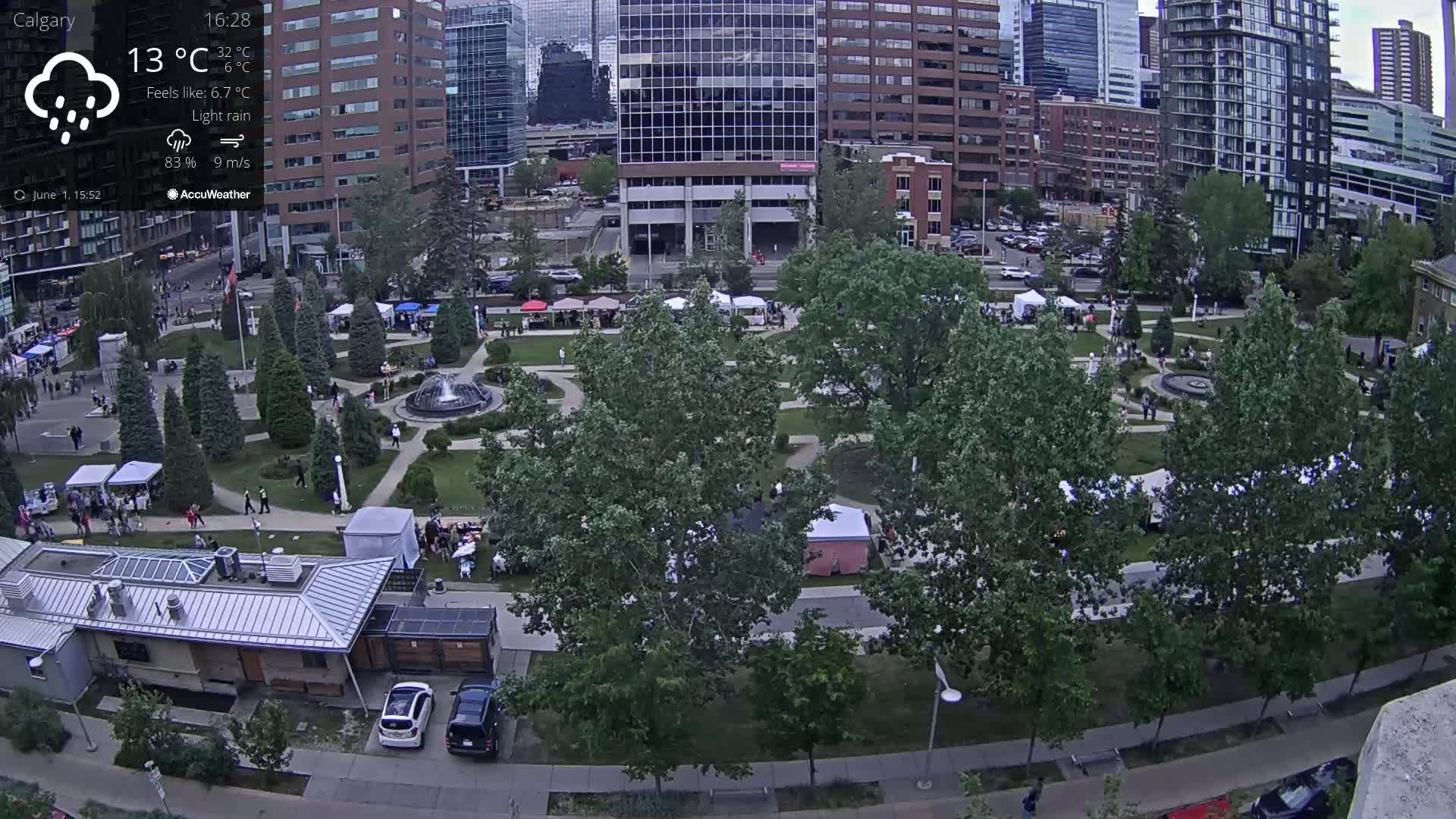 An aerial view shows a park with a fountain and numerous people amidst market stalls under a light rain.