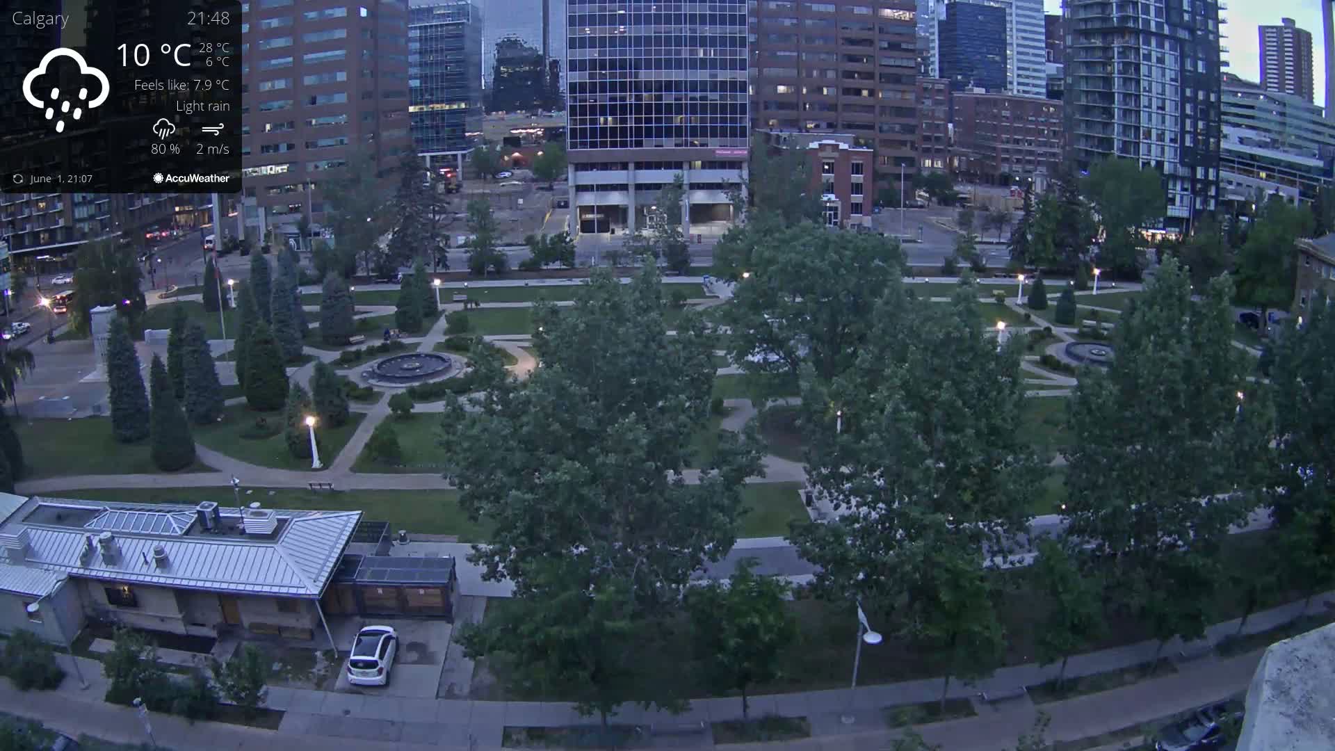 An aerial view shows a city park with a fountain at twilight under light rain, surrounded by tall buildings and trees.
