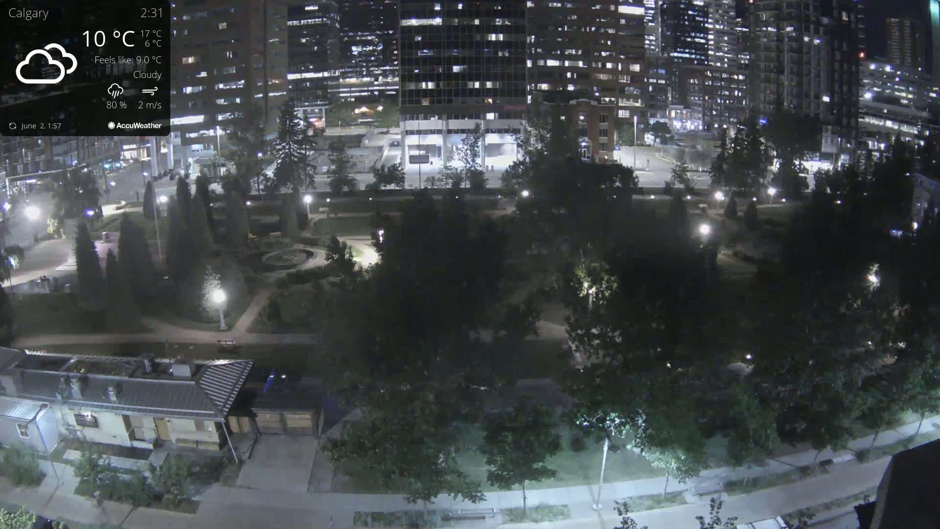 A nighttime, cloudy view of a city park bordered by illuminated buildings and pathways.