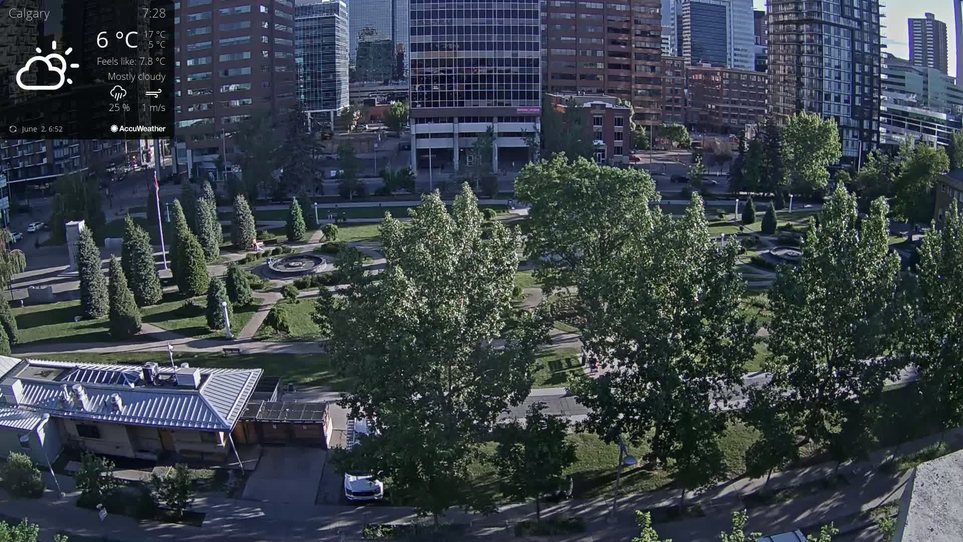 A mostly cloudy city park with a fountain, surrounded by tall buildings, is seen from an elevated view.