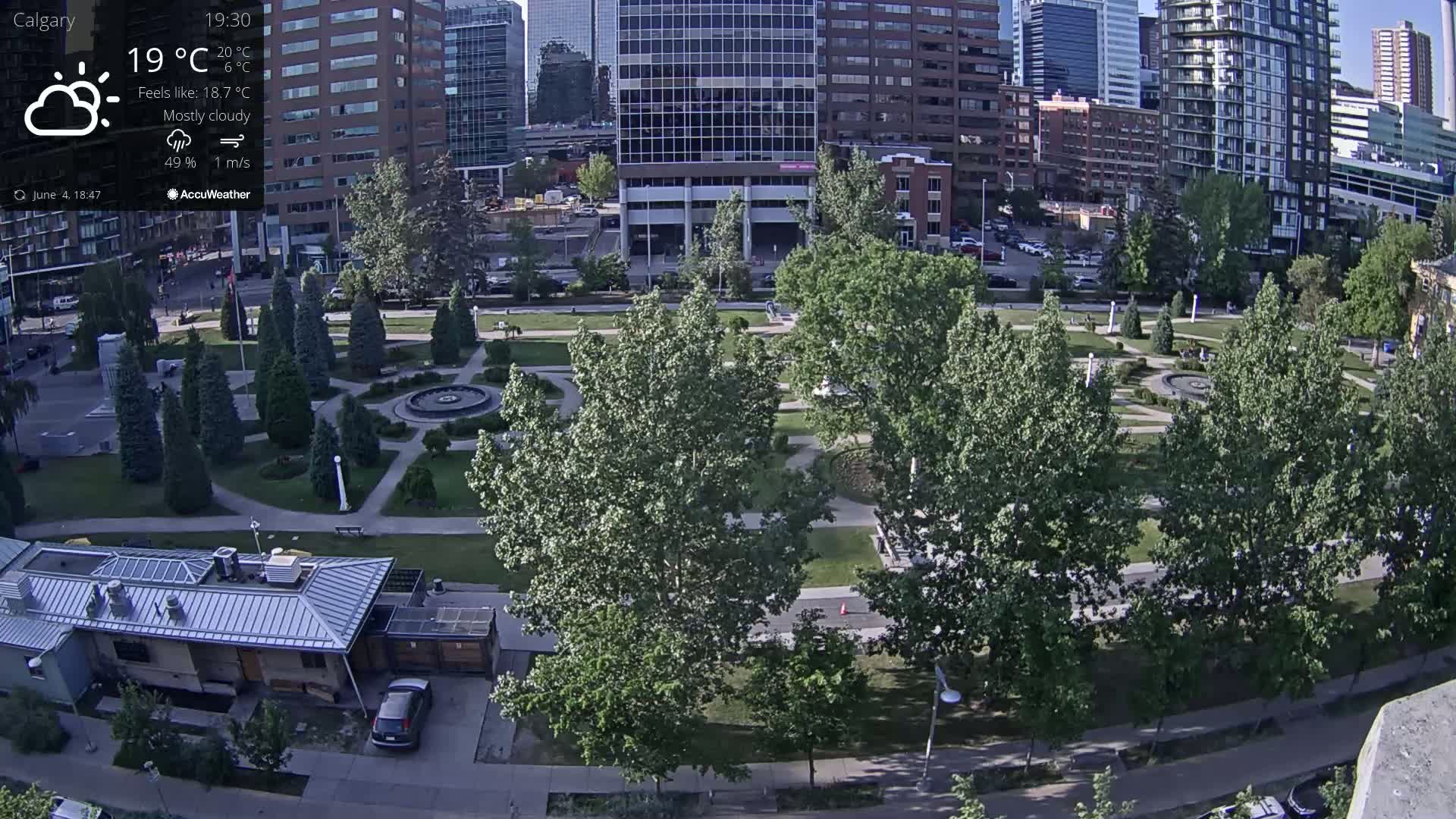 A mostly cloudy, 19°C day shows a city park with a fountain, surrounded by green trees and tall buildings.