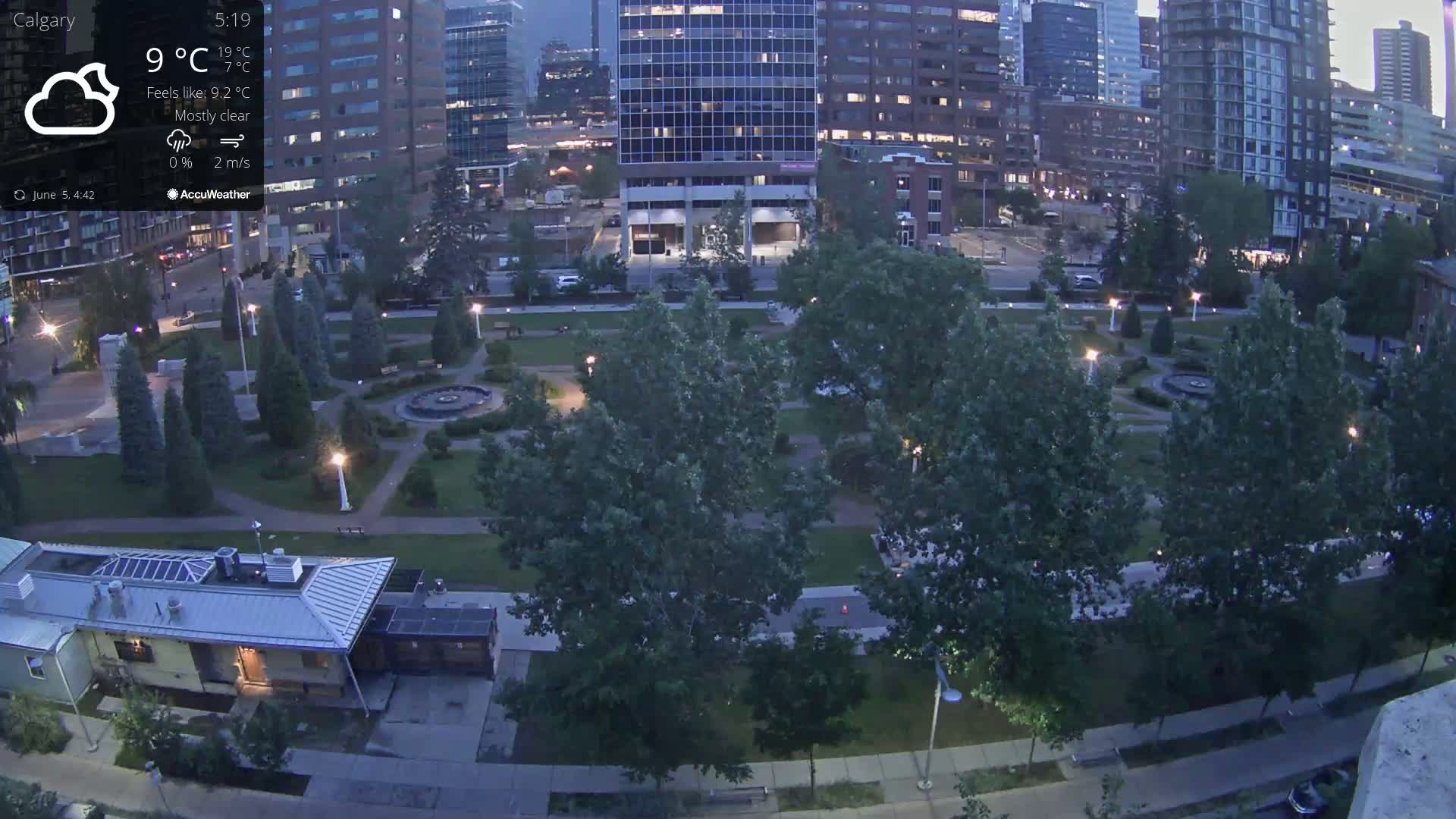 A mostly clear evening scene shows a city park with trees and pathways, bordered by tall buildings under a mostly clear sky.