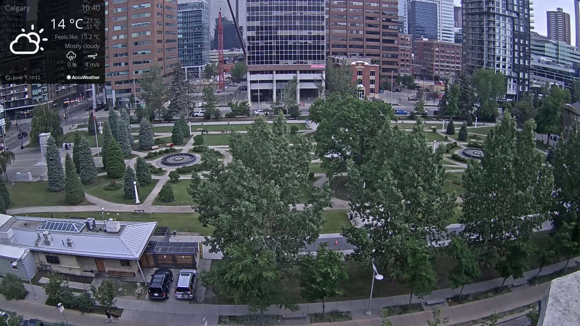 A mostly cloudy cityscape features a park with paved pathways, lush green lawns, and numerous trees surrounding a central fountain, with several city buildings in the background.
